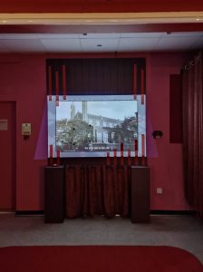 The setting is a dimmed room. The walls are pink, on the floor - at the bottom of the photo - an edge of a red carpet peeks. At the centre is an installation with projected video.