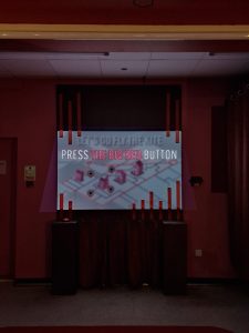 Photo of a dimmed room with deep burgundy hue. At the centre of the image is a projected content of a map, blurred, with clear large font reading Press the big red button.