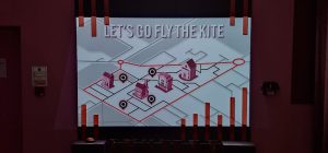 A close up of a projected content on a wall. The room is dim and the colours of the walls and the installation almost blend together into dark burgundy. The projected content is a map on a grey background. The area of interest is highlighted in deep pink and four buildings are put at key locations with location pins positioned next to them.